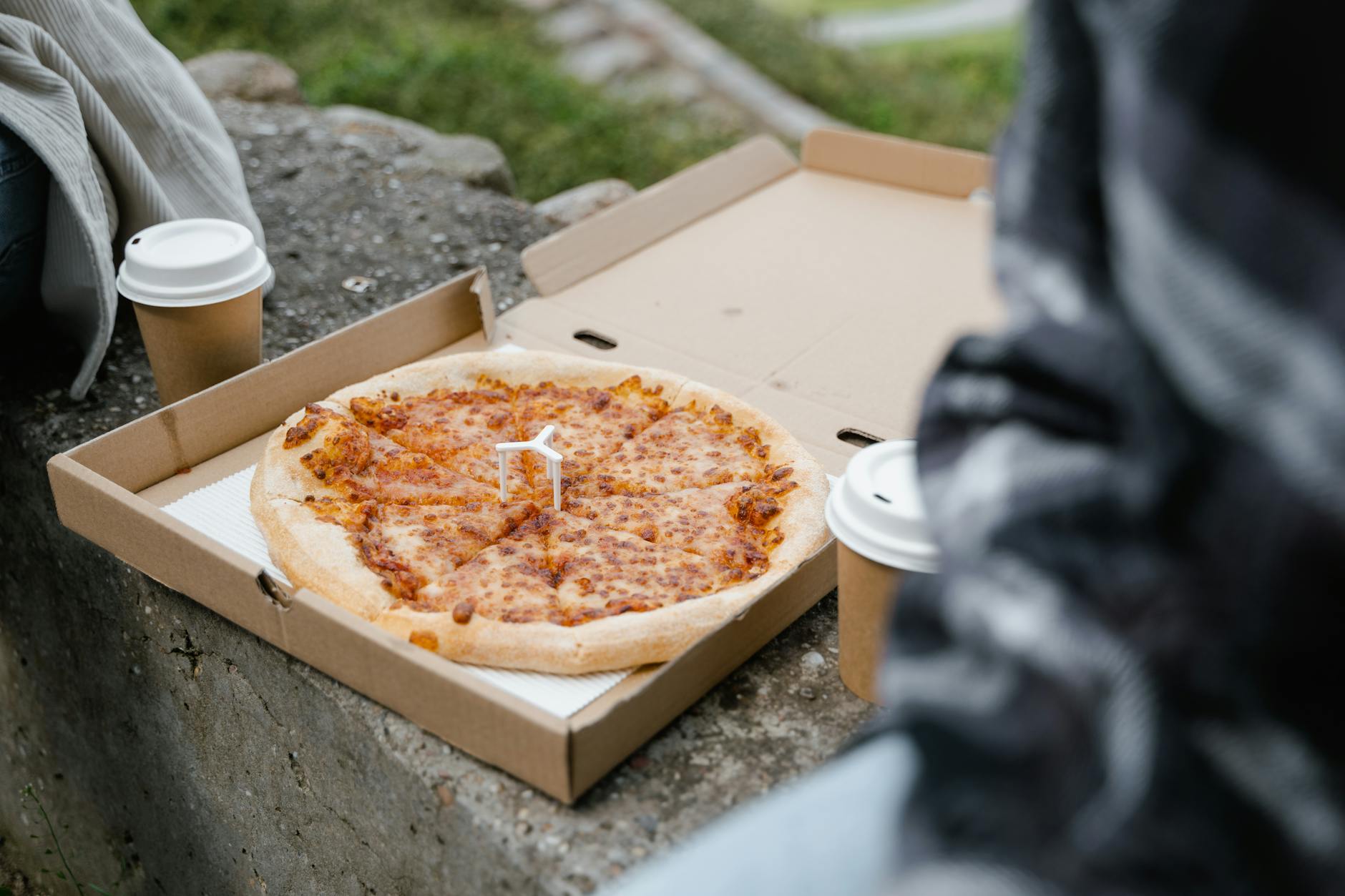 Otwarty karton z pizzą i papierowe kubki kawy na kamiennej ławce