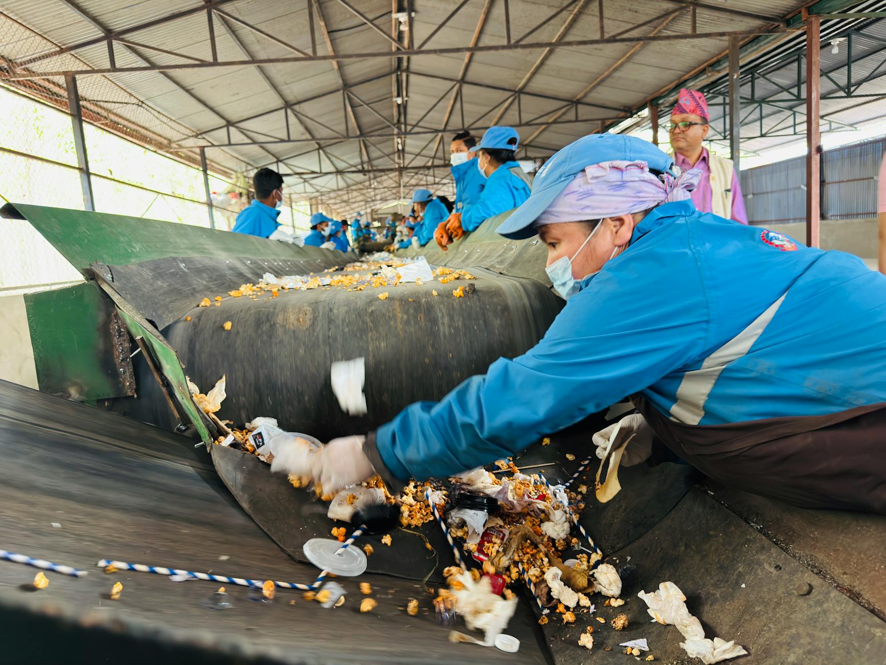 Pracownicy w niebieskich uniformach sortują odpady do recyklingu na taśmie